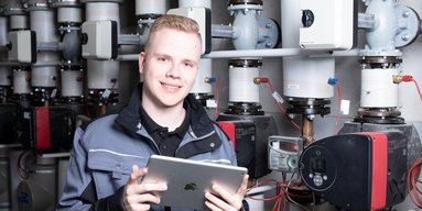 Ein junger Anlagenmechaniker vor einer Heizungsanlage, der ein Tablet hält 