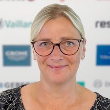 Professionelle Porträtaufnahme einer lächelnden Frau Eine Frau mit blonden Haaren und Brille lächelt freundlich vor einem verschwommenen Hintergrund mit Logos.