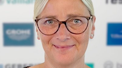 Eine Frau mit blonden Haaren und Brille lächelt freundlich vor einem verschwommenen Hintergrund mit Logos.