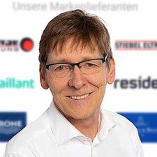 Professionelles Porträt eines freundlichen Mannes mit Brille Ein lächelnder Mann mit Brille trägt ein weißes Hemd, vor einem verschwommenen Hintergrund mit Markenlogos.