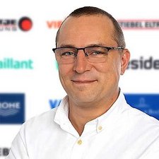 Professionelles Porträt eines lächelnden Mannes mit Brille Ein lächelnder Mann mit Brille trägt ein weißes Hemd und steht vor einem Hintergrund mit verschiedenen Logos.
