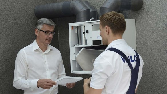 Draußen stehen zwei Männer, der Linke ist ein Kunde und der Rechte ein Mechatroniker. der Kunde hält einen Filter von der Lüftungsanlage in der Hand. Der Mechatroniker wartet die Anlage.